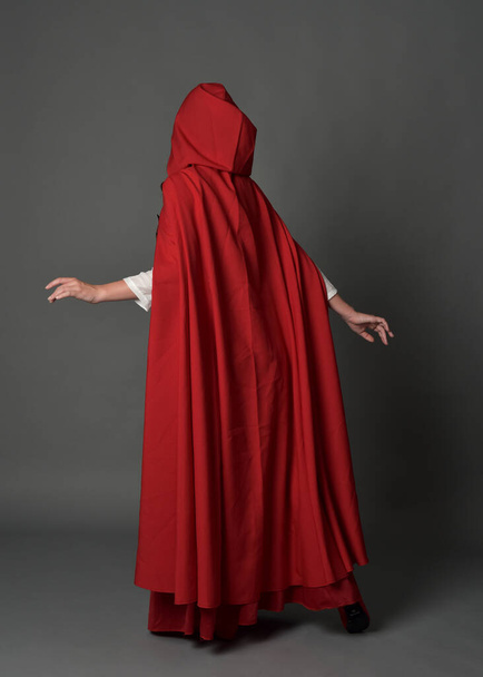 Full length portrait of woman wearing red medieval fantasy costume, flowing hooded cloak. Standing pose in backview, gestural hand poses, walking away from camera isolated on grey studio background. - Photo, Image