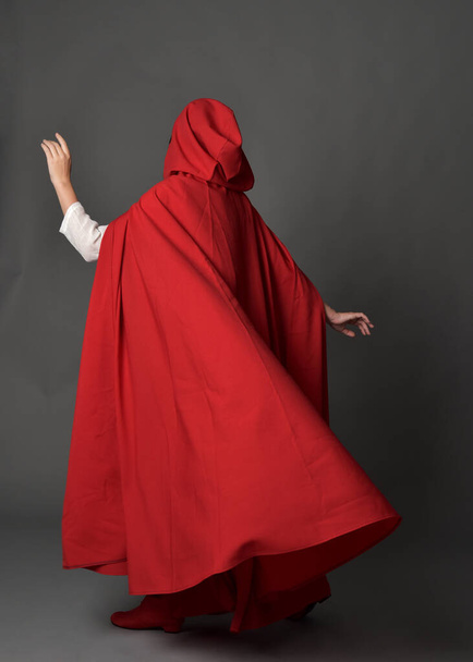Full length portrait of woman wearing red medieval fantasy costume, flowing hooded cloak. Standing pose in backview, gestural hand poses, walking away from camera isolated on grey studio background. - Фото, изображение