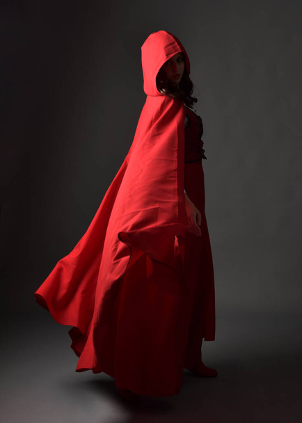 Täyspitkä muotokuva nainen yllään punainen keskiaikainen fantasia puku, virtaava hupullinen viitta. Seisova poseeraa taustakuvassa, eleellinen käsi aiheuttaa, kävely pois kameran eristetty harmaa studio tausta. - Valokuva, kuva