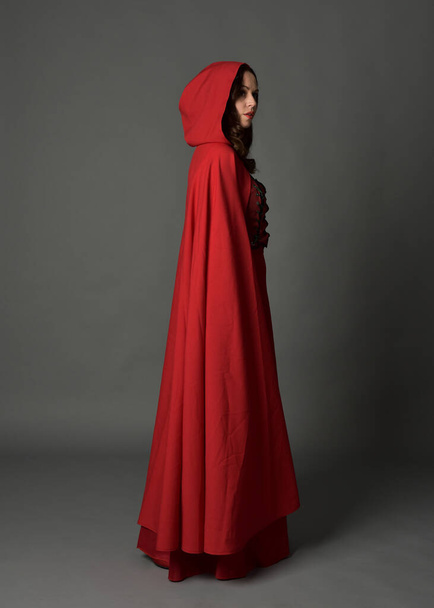 Full length portrait of woman wearing red medieval fantasy costume, flowing hooded cloak. Standing pose side profile, gestural hand poses, walking away from camera isolated on grey studio background. - Photo, image