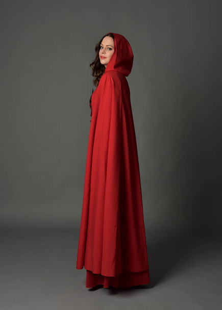 Повнометражний портрет жінки в червоному середньовічному фентезійному костюмі, що тече з капюшоном плащ. Стоїть профіль сторони позу, жестова рука позує, йде від камери ізольовано на сірому студійному фоні
. - Фото, зображення