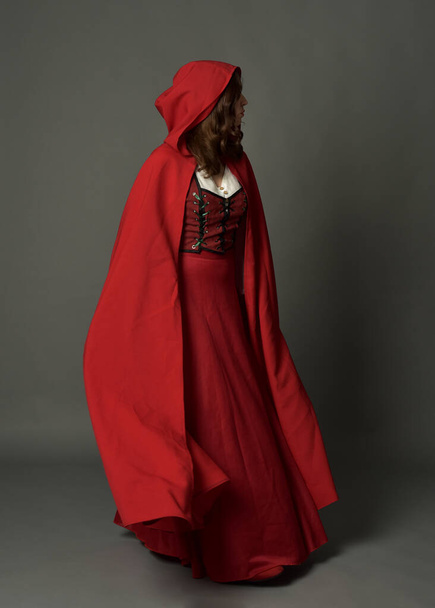 Retrato de comprimento total de mulher vestindo traje de fantasia medieval vermelho, manto com capuz fluindo. Perfil lateral da pose em pé, poses gestuais da mão, afastando-se da câmera isolada no fundo do estúdio cinza. - Foto, Imagem
