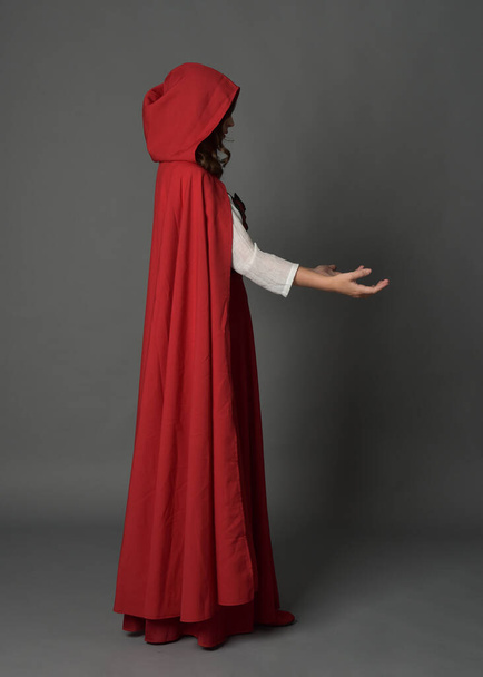 Полнометражный портрет женщины в красном средневековом сказочном костюме в капюшоне. Стоячая поза боковой профиль, жесты руки позы, уход от камеры изолированы на сером фоне студии. - Фото, изображение