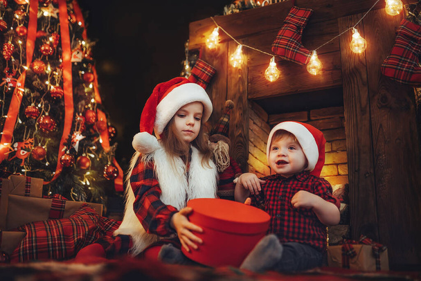 Children in Santas hats open giftbox near fireplace and christmas tree. New Year concept. Christmas time  - Φωτογραφία, εικόνα
