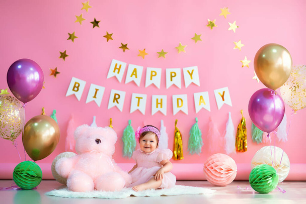 Cute baby girl sits near paper balls and colorful air balloons with pink teddy in pink holiday dress on pink background with letters . happy birthday concept - Foto, Imagem