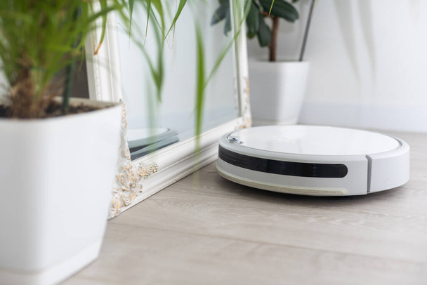 A white robot vacuum cleaner vacuums along the skirting Board near a large window through which the bright sun shines. close-up, beige colors, small depth of field. - Photo, Image