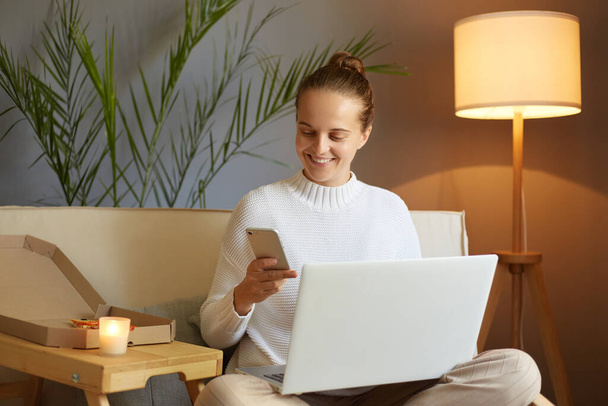 Indoor shot of attractive happy woman using multiple devices while sitting on sofa, looking at mobile phone display, being in good mood, working at home. - Foto, afbeelding