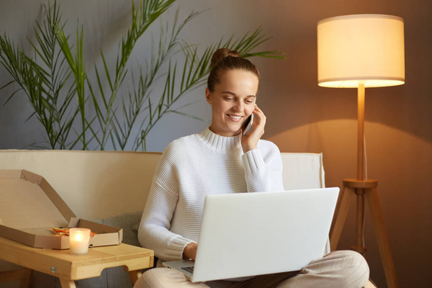Indoor shot of smiling attractive positive Caucasian woman wearing white sweater sitting on sofa and working on laptop in home interior, talking on smart phone. - Foto, Bild