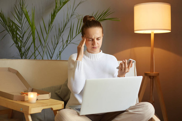 Horizontale Aufnahme einer müden, kranken Frau mit weißem Pullover, die auf dem Sofa sitzt und an Laptop und Handy arbeitet, Kopfschmerzen hat und viel Arbeit hat, hält die Hand an der Schläfe. - Foto, Bild