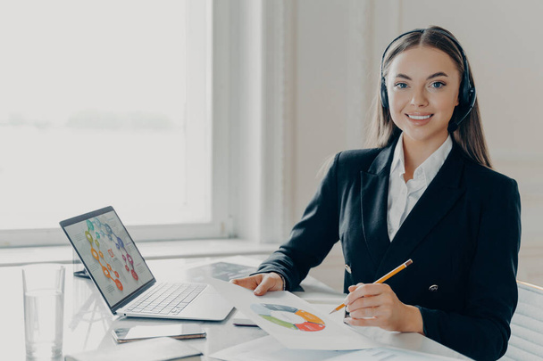 Young happy female office worker or employee in formal black suit preparing financial report or business presentation, sitting at workplace in offce. Smiling businesswoman analyzing data on laptop - Foto, Imagen