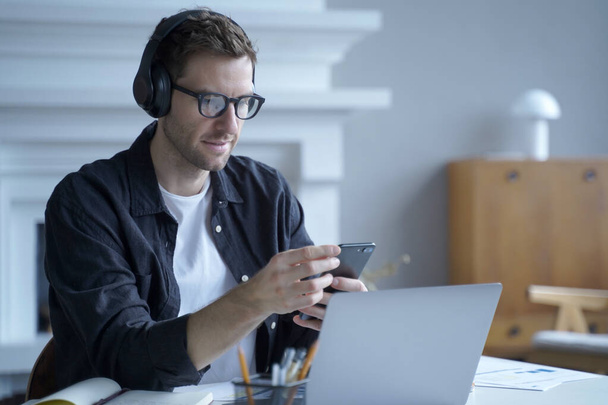 Serious austrian guy freelancer in headphones using phone at workplace, sitting at desk at home office working on laptop and at same time responding on important email or sms on smartphone in hands - Фото, изображение