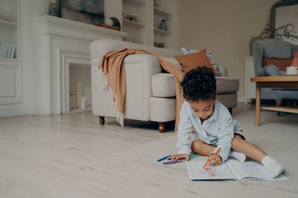 Μικρή μικτή φυλή παιδί σχέδιο αγόρι στο βιβλίο ζωγραφικής με πολλά διαφορετικά στυλό άκρη τσόχα, ενώ κάθεται μόνη της στο πάτωμα στη μέση του σαλονιού στο σπίτι. Παιδικές δραστηριότητες αναψυχής και ανάπτυξης - Φωτογραφία, εικόνα