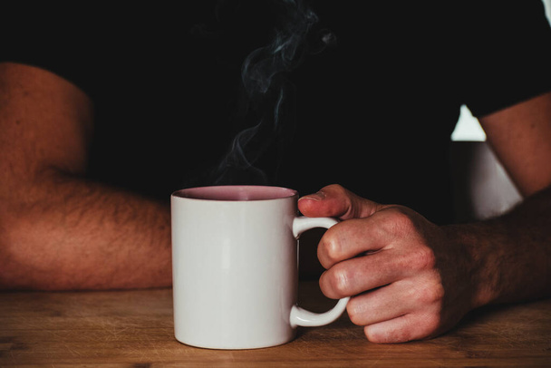 man holding steaming mug on a wooden table - Фото, зображення