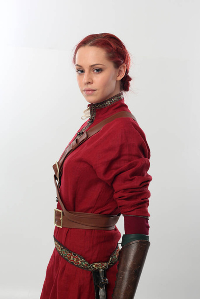 πορτρέτο της όμορφης γυναίκας φορώντας ένα κόκκινο μεσαιωνικό κοστούμι πολεμιστή φαντασίας με δερμάτινη πανοπλία, κρατώντας όπλα. Μόνιμη στάση απομονωμένη σε φόντο στούντιο. - Φωτογραφία, εικόνα