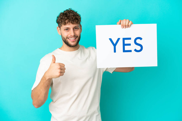 Молодий красивий кавказький чоловік ізольований на синьому фоні, тримаючи плакат з текстом YES з великим пальцем вгору
 - Фото, зображення