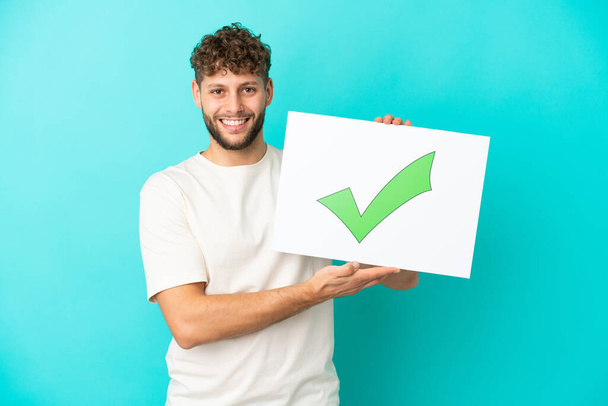 Young handsome caucasian man isolated on blue background holding a placard with text Green check mark icon with happy expression - Photo, Image