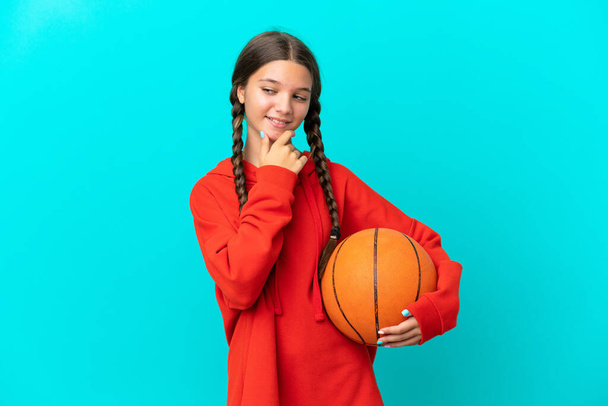 Pequeña chica caucásica jugando baloncesto aislado sobre fondo azul mirando a un lado y sonriendo - Foto, imagen