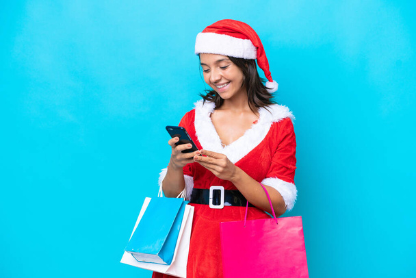 Mujer hispana joven vestida como mamá Noel aislada sobre fondo azul sosteniendo bolsas de la compra y escribiendo un mensaje con su teléfono celular a un amigo - Foto, imagen