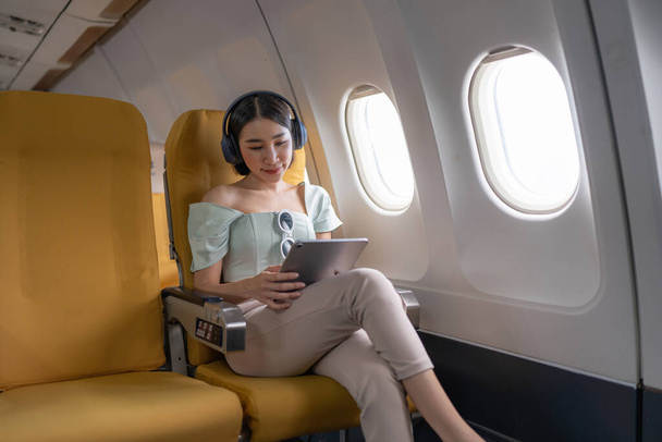楽しいアジアの女性は飛行機に座って、旅行に行く間タブレットを使用しています. - 写真・画像