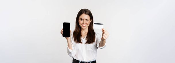Portrait of female model showing credit card with smartphone screen, recommending application, standing over white background. - Fotó, kép
