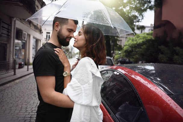 Young couple with umbrella enjoying time together under rain on city street, space for text - Valokuva, kuva