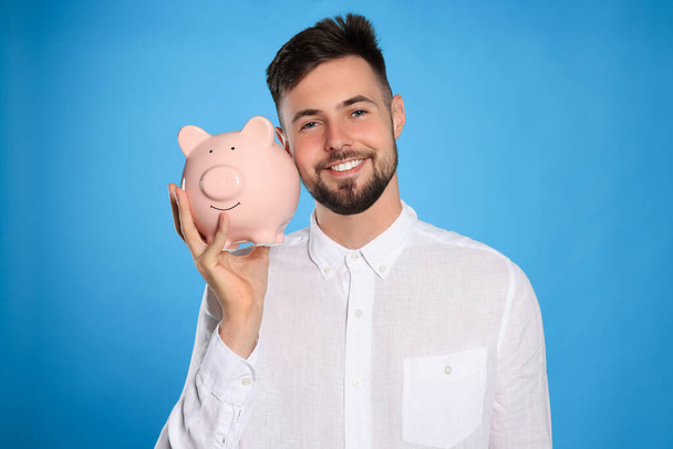 Happy young man with piggy bank on light blue background - Фото, изображение