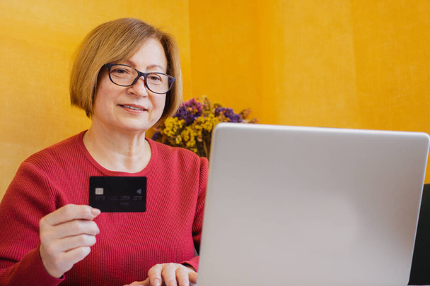 Boldog érett nő szemüvegben, aki bankkártyával nézi a laptopot, internetes fizetést teljesít, online vásárol, szállodát foglal, taxit rendel alkalmazáson keresztül, bent ül. Nyugdíjazás és online banki szolgáltatások. - Fotó, kép
