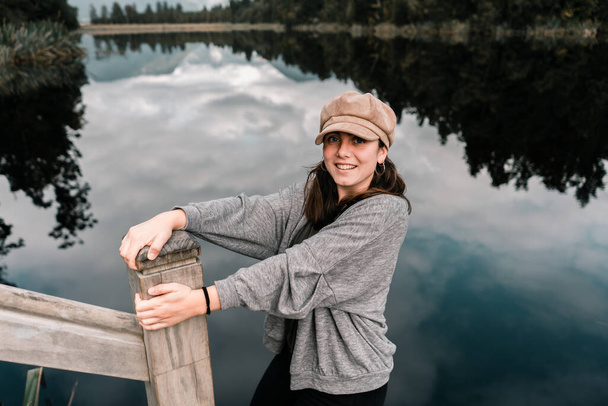 caucasian girl in nice brown hat gray jacket and black pants having fun happy with outstretched arms tightly holding on to sturdy wooden post on shore of calm lake in green forest, new zealand - Photo, Image