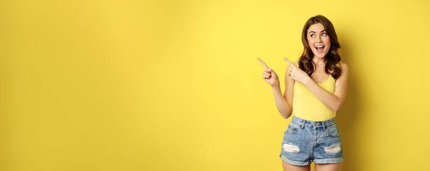 Şık bir yaz kızının portresi, yol gösteren esmer kadın, sol parmakları gösteren, reklam yapan, sarı arka planda duran. Boşluğu kopyala - Fotoğraf, Görsel