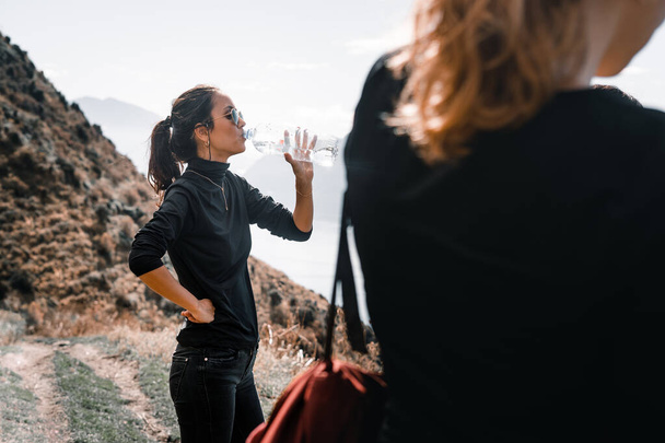 Кавказская девушка в черной футболке солнцезащитные очки черные брюки правая рука на талии пить пресную воду с левой рукой из пластиковой бутылки на обочине дороги - концепция путешествия - Фото, изображение