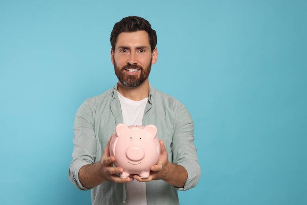 Happy man with ceramic piggy bank on light blue background, space for text - Fotoğraf, Görsel