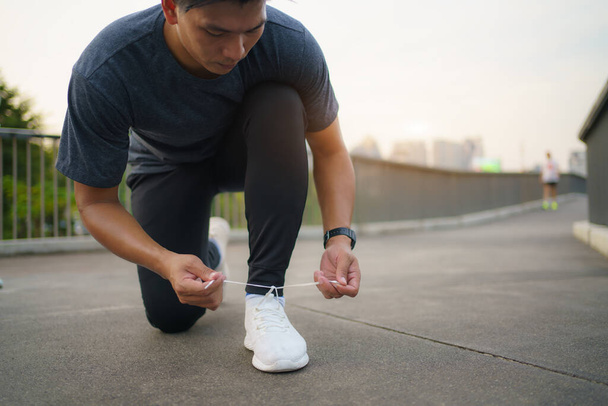 Close-up of Asian man tying shoelaces on white sneakers. Male person squatting on sidewalk. Sunny morning or day. Male going on footpath. Sport and healthy lifestyle concept - Fotografie, Obrázek