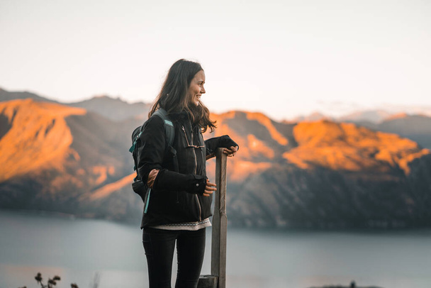 caucasian girl standing leaning on wooden post wearing black coat black pants glasses around her neck backpack on her back in profile happy calm and relaxed on mountain summit at sunset, roys peak - Foto, imagen