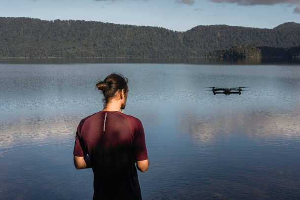 long-haired caucasian boy with a drone in the air controlled by him on the shore of the calm lake and near the impressive forest full of trees in the middle of nature, new zealand glaciers - Nature - Photo, Image
