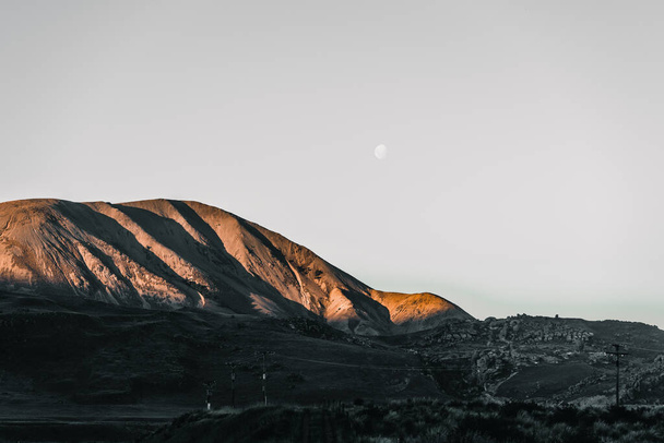 rocky mountains in sunset sunlight in a sad and desolate landscape with very little vegetation power poles and wires under the waning moon in a gray sky, arthur pass, new zealand - Travel Concept - Photo, image