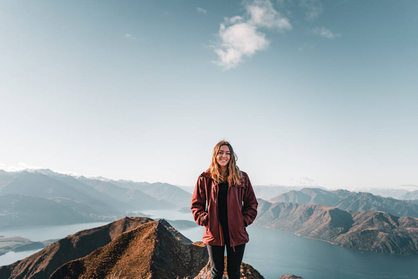 blondýny běloška dívka na sobě hnědé sako černé kalhoty a ruce v kapsách s úsměvem na fotoaparátu klidné a uvolněné z vrcholu hory s výhledem na velké modré jezero a ohromující hory, roys vrchol, nové - Fotografie, Obrázek