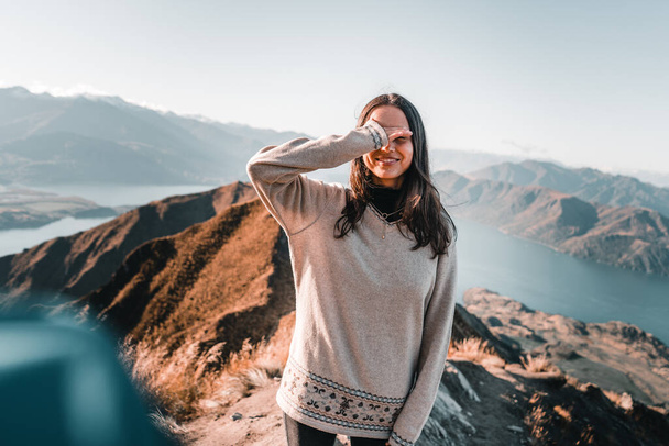 morena chica caucásica con el pelo largo gris suéter pantalones negros con la mano derecha que cubre su cara feliz desde la cima de la montaña en un paisaje tranquilo en medio de la naturaleza, roys pico, Nueva Zelanda - Foto, imagen