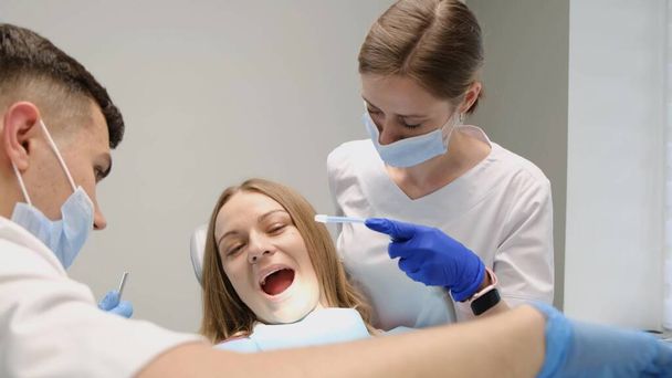 Современная стоматология. Молодой врач и ассистент лечат зубы молодой женщины. Безболезненное лечение зубов. Своевременное лечение зубов. - Фото, изображение