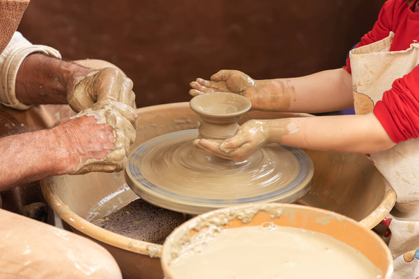 close-up in horizontaal zicht van de handen van een pottenbakker en een meisje, terwijl de pottenbakker het meisje leert om een kleibeker te modelleren. leerlingwezen in de traditionele beroepen - Foto, afbeelding