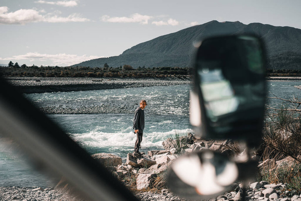 caucasiano menino desgrenhado fotógrafo de pé olhando para a câmera de pé em uma rocha junto ao rio com a van estacionada perto de montanhas e natureza, rio taramakau, Nova Zelândia conceito de viagem - Foto, Imagem