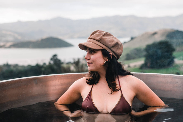 blue-eyed caucasian young woman looking to the side wearing a swimsuit and a brown cap inside the small round jacuzzi with a view of the mountains and french farm bay, te wepu pods akaroa, new zealand - Foto, Imagem