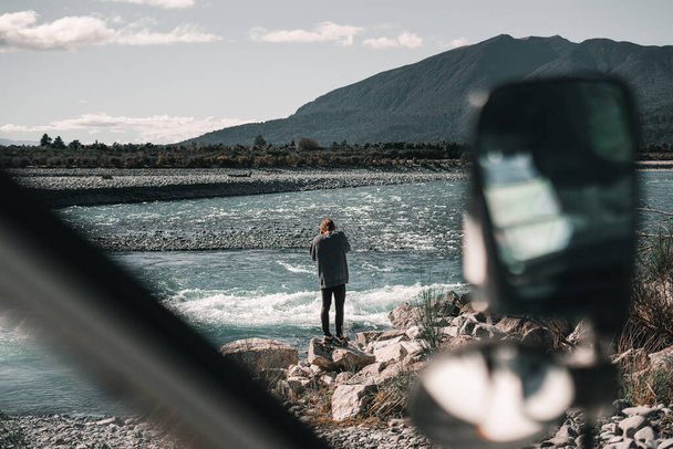 menino quacassiano em pé sobre uma rocha na margem do rio grande e solitário fotografando as ondas formadas pela corrente da água perto das montanhas na natureza, rio taramakau, nova zelândia - Viajar - Foto, Imagem