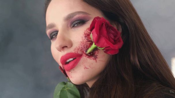 Retrato de una niña sosteniendo una rosa roja en su boca. Maquillaje para Halloween, rosa flor en la boca. Contra el fondo del humo - Foto, imagen