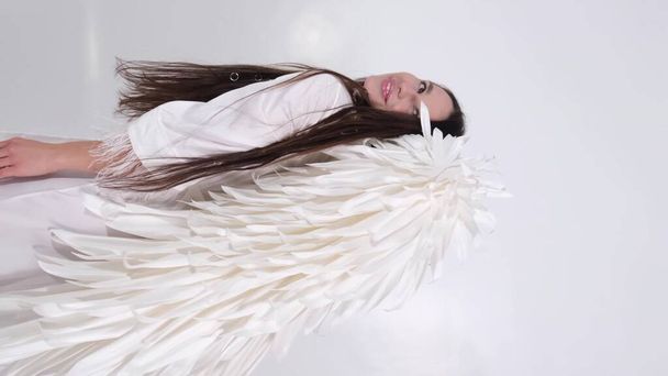 Vidéo verticale, belle brune aux ailes blanches sur fond blanc. Une fille en costume d'ange. Vidéo 4k de haute qualité - Photo, image