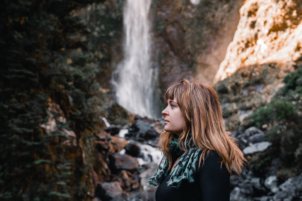 caucasian girl with black t-shirt and thin green scarf wrapped around her neck looking serious and sad in profile with the waterfall and the river behind her at sunset, matheson lake, new zealand - - Photo, Image