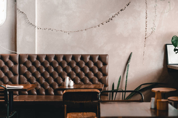 Sessiz ve dost canlısı bir kahve dükkanındaki ahşap masaları, Mylk Bar 'ı, Yeni Zelanda' daki yaşam tarzı konseptini, eski moda rahat deri koltuğun üzerinde asılı şirin yapraklı bitkilerle dolu gri bir duvar. - Fotoğraf, Görsel