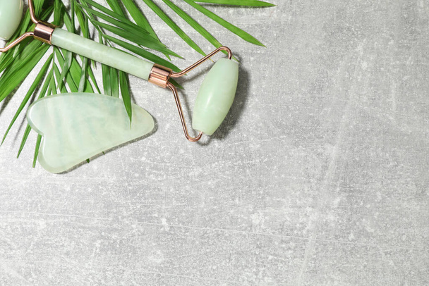 Quartz gua sha tool, face roller and green leaves on grey background, flat lay. Space for text - 写真・画像