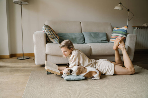 Attractive blonde woman enjoying leisure time reading novel home with small friend dog Jack Russell terrier sleeping. Relaxed moments chilling at home with your pet. Slow life atmosphere vibe - Foto, Bild