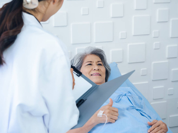 笑顔アジアのシニア女性灰色の髪の患者がベッドに横たわって、若い女性医師が訪問しながら回復し、病院の部屋で症状や検査のメモや医療文書を説明します.  - 写真・画像