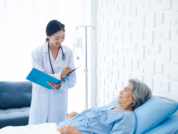 Lächelnde asiatische Seniorin mit grauen Haaren, die auf dem Bett liegt und sich erholt, während junge Ärztinnen zu Besuch kommen, um die Symptome und Untersuchungsnotizen und medizinischen Unterlagen im Krankenhauszimmer zu erklären.  - Foto, Bild